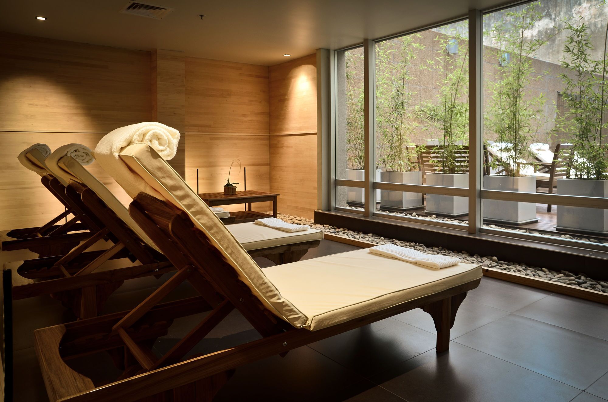 A room with two wooden massage tables, a small table with a plant, and a view of a bamboo garden through large windows.