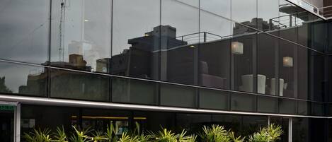 Un edificio moderno con fachada de vidrio reflectante, una piscina con escalera metálica y macetas alineadas.