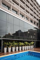 Un edificio moderno con fachada de vidrio reflectante, una piscina con escalera metálica y macetas alineadas.