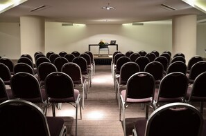 Un salón de conferencias con filas de sillas orientadas hacia un escenario, donde hay una computadora portátil y una planta.