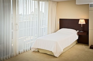 Habitación de hotel con una cama, una mesita de noche, una lámpara y vistas a través de cortinas translúcidas.