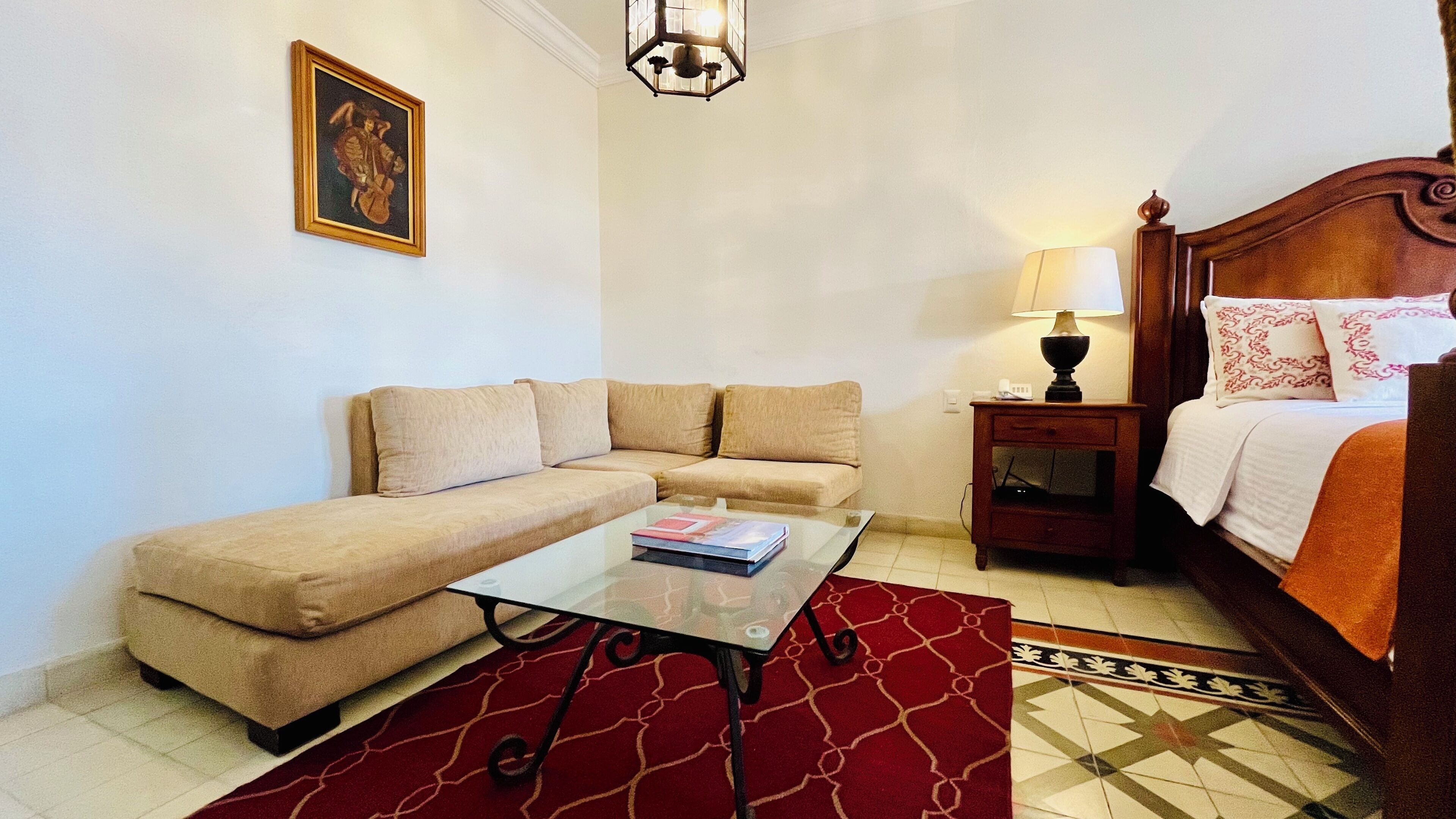 Una habitación con sofá color beige, una mesa de centro de vidrio, una mesita de noche de madera con lámpara, cama con ropa blanca y almohadas rojas, y una alfombra estampada.
