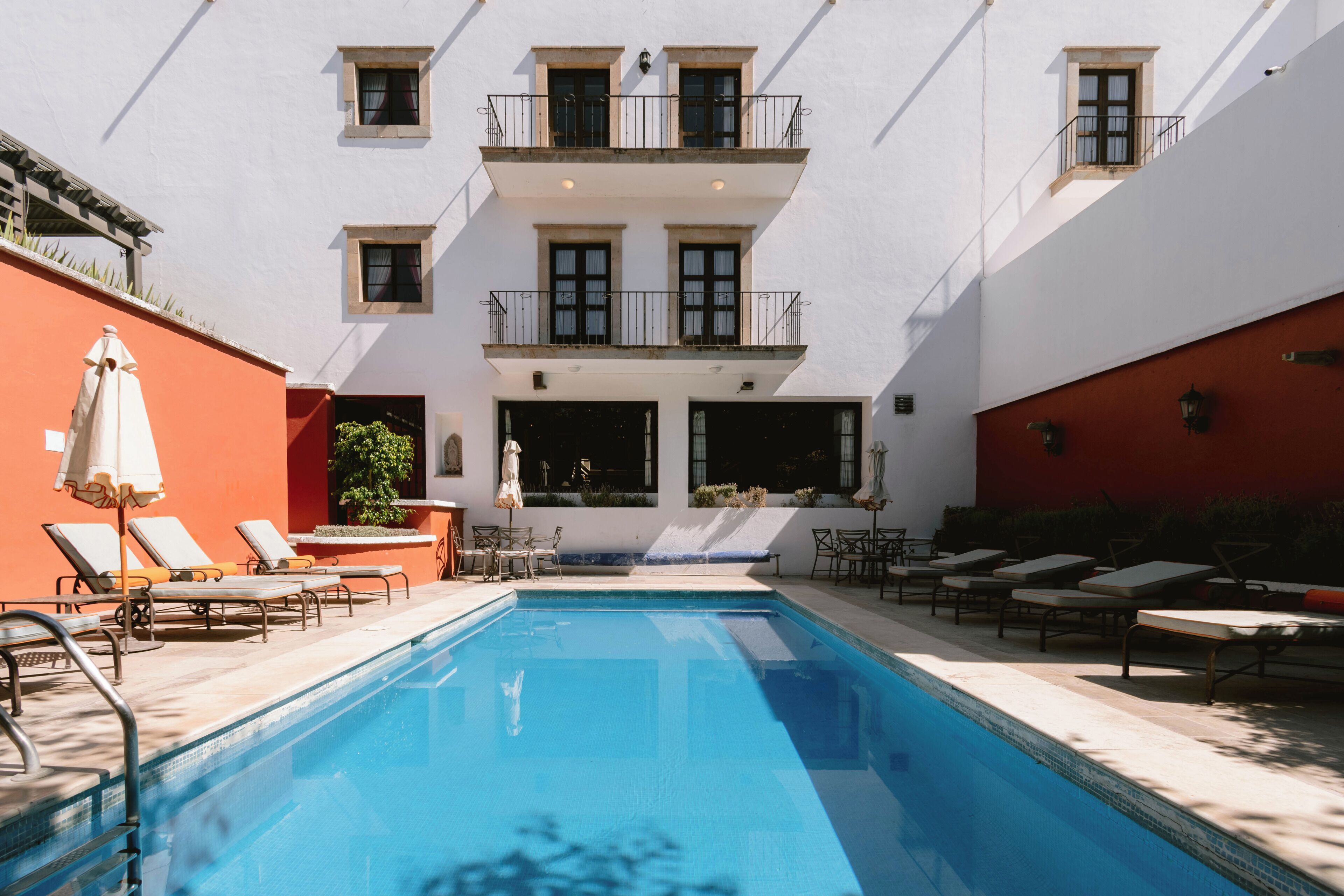 A swimming pool with sun loungers and a small table, surrounded by a white building with balconies.