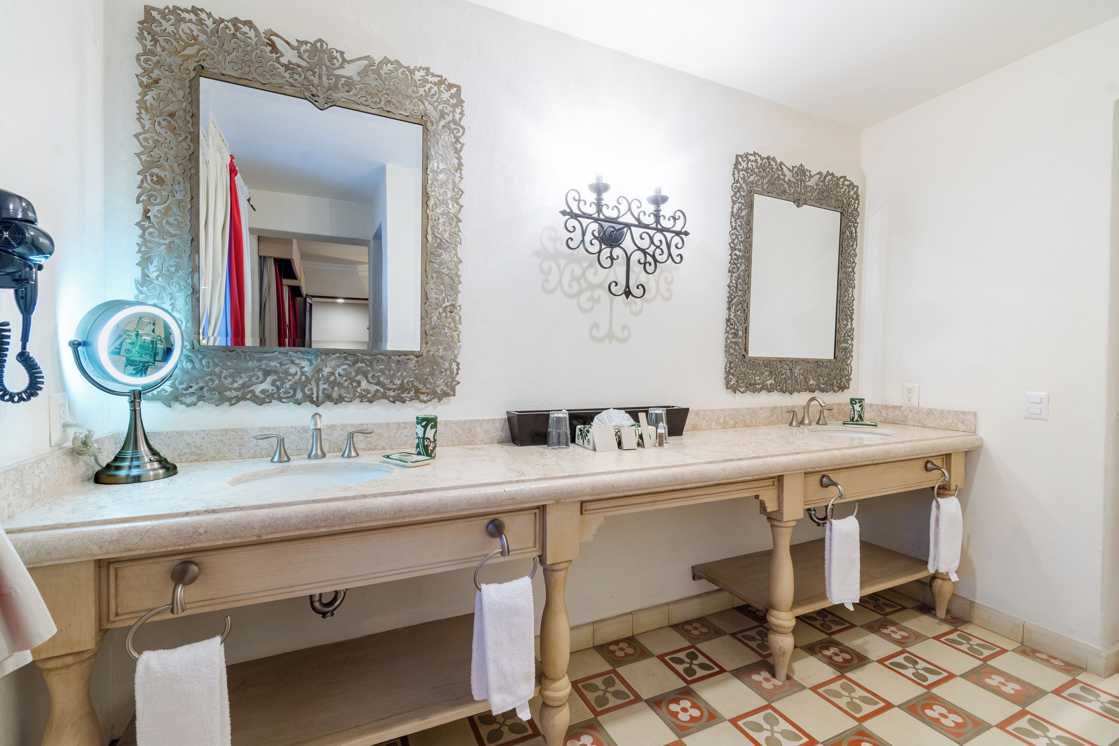 A bathroom with two sinks, ornate mirrors, a vanity with drawers, and a patterned floor.