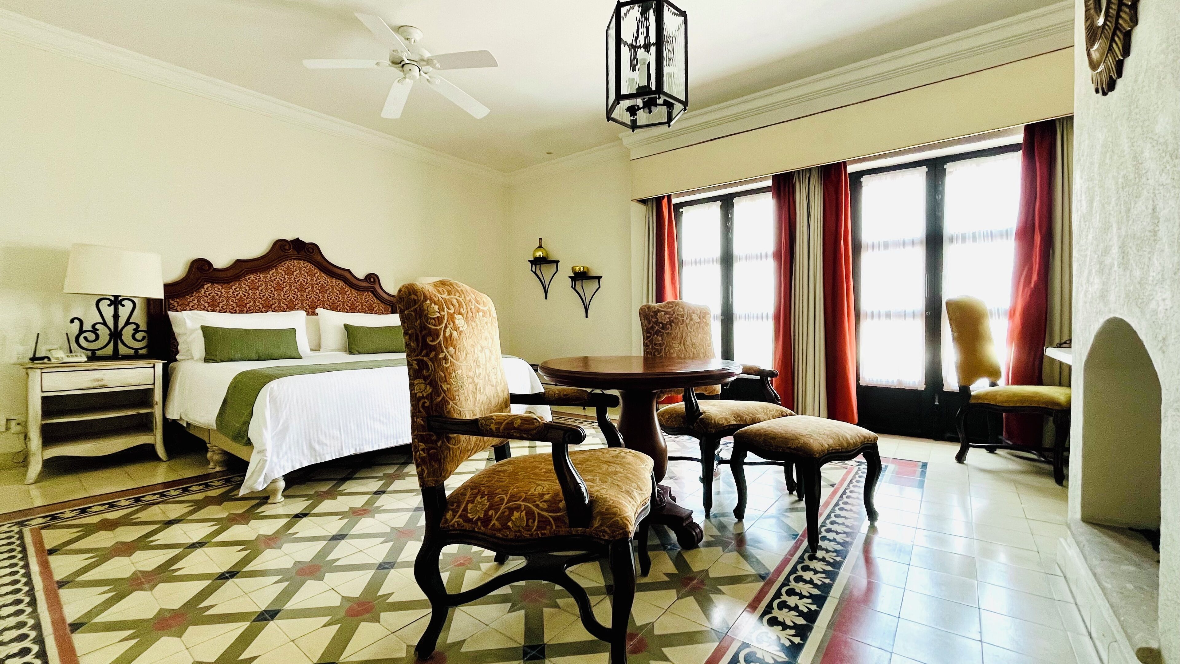 A hotel room with a bed, two armchairs, a table, a lamp, and a chandelier.