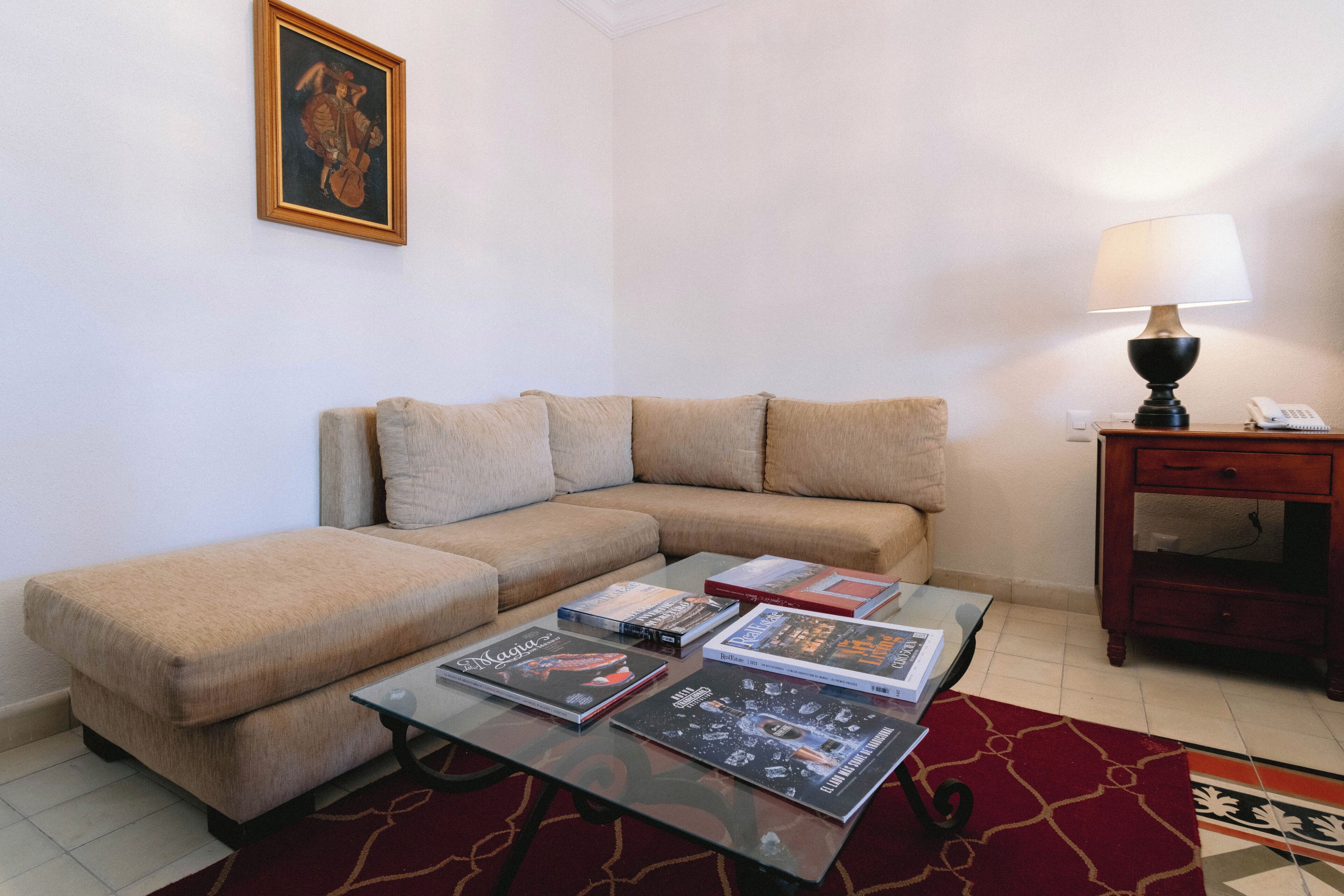 A living room with a beige sectional sofa, a glass coffee table with magazines, a lamp, and a small wooden side table with a phone.