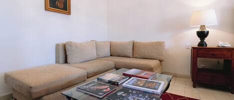 A living room with a beige sectional sofa, a glass coffee table with magazines, a lamp, and a small wooden side table with a phone.