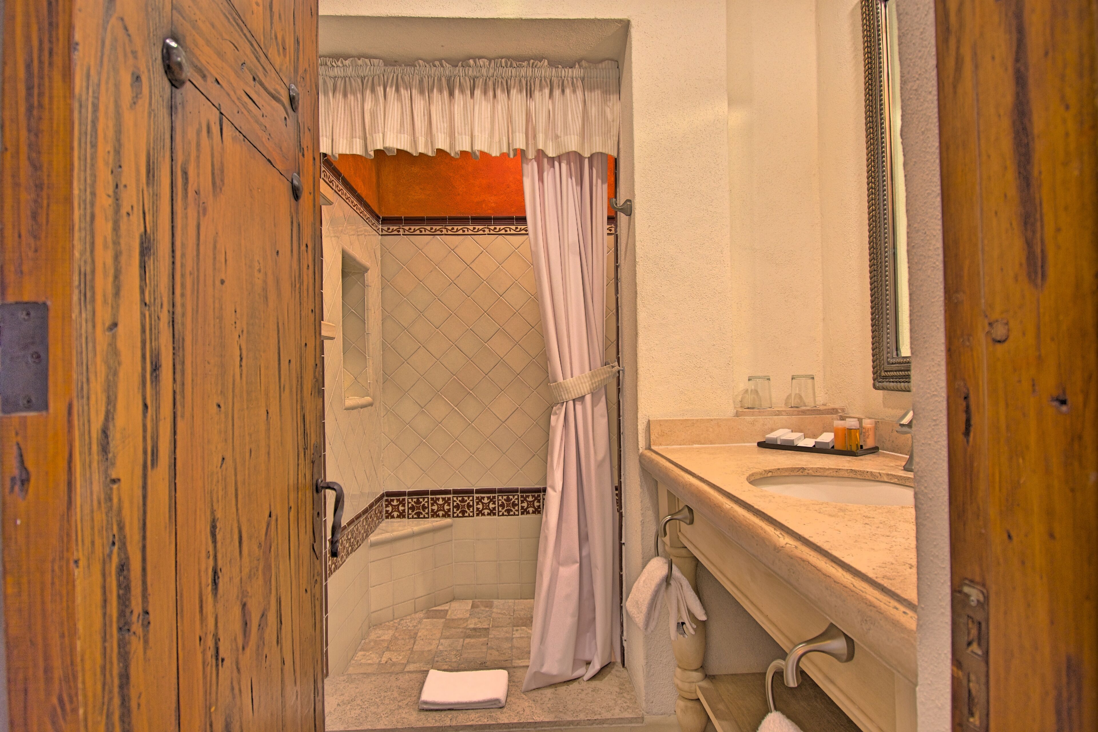 Une salle de bain avec une porte en bois, une douche avec un rideau, un lavabo avec des articles de toilette et un miroir.