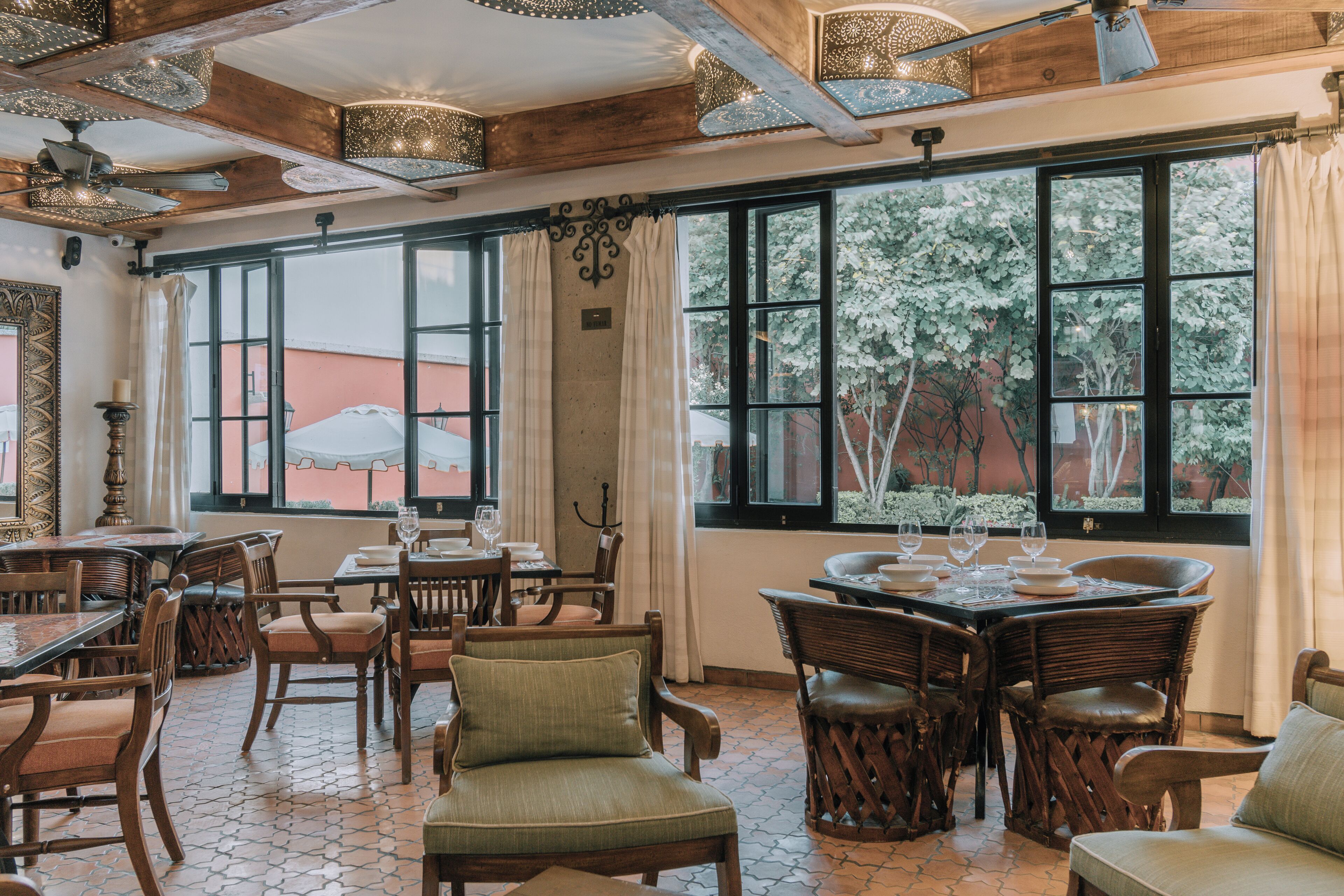 Un comedor con mesas y sillas de madera, ventanas amplias con cortinas y suelo de baldosas.