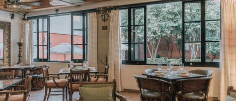 Un comedor con mesas y sillas de madera, ventanas amplias con cortinas y suelo de baldosas.