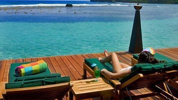 Eine Holzterrasse mit Liegestühlen und einem Tisch, mit Blick auf das türkisfarbene Meer und eine kleine Insel.