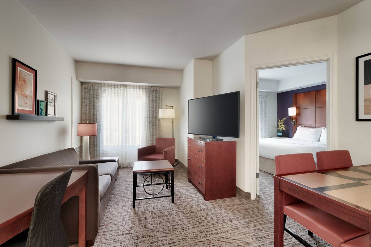 A hotel room with a sofa, a small table, a TV on a wooden cabinet, a dining table with chairs, and a bed in the adjacent room.