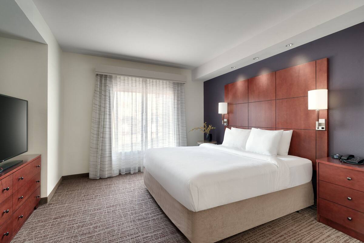 A hotel room with a large bed, a TV on a dresser, a nightstand with a lamp, and a window with curtains.
