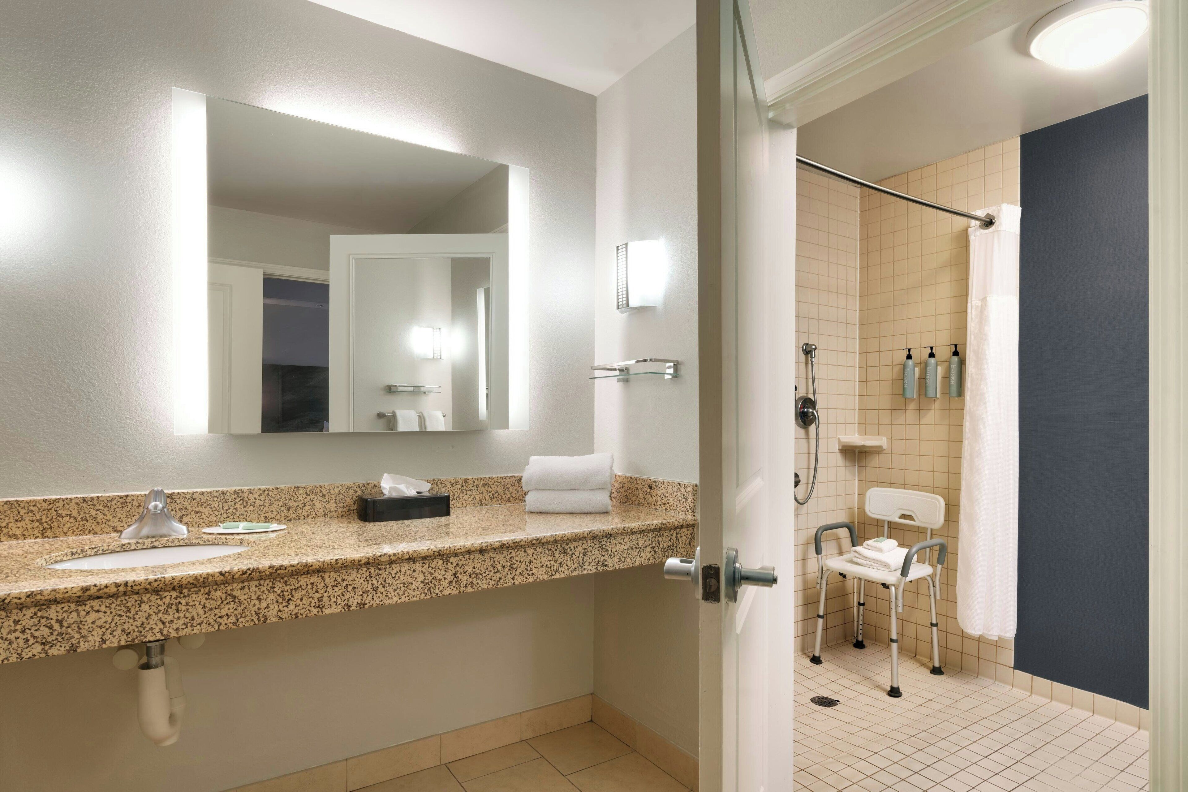 A bathroom with a granite countertop, a mirror, a towel rack, a shower area with a grab bar, and a white chair.