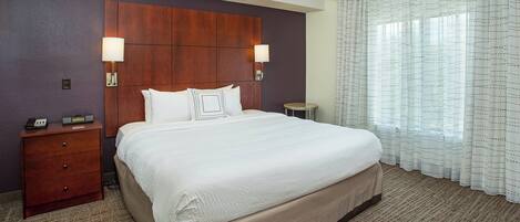 Una habitación de hotel con una cama grande, dos lámparas de noche, un cabecero de madera, una mesita de noche con un teléfono y una ventana con cortinas traslúcidas.