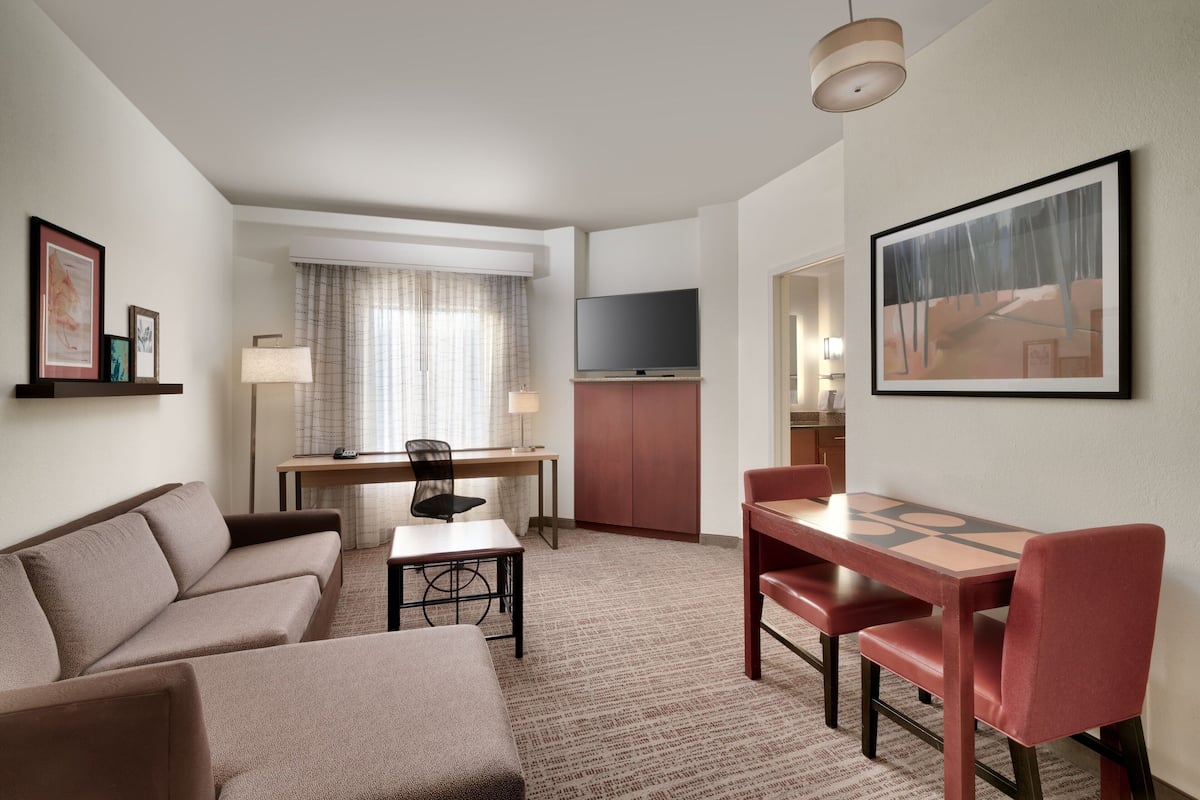A modern hotel room with a sofa, a dining table with chairs, a TV, and artwork on the walls.