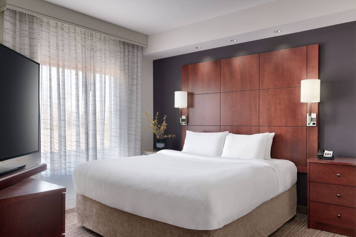 A modern hotel room with a large bed, a flat-screen TV, a wooden headboard, and a bedside table with a lamp.