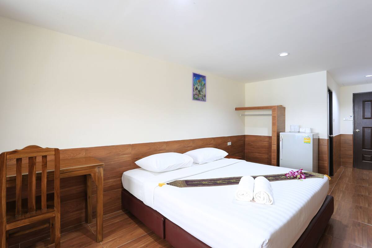 A hotel room with a wooden bed, white linens, a small wooden table, a refrigerator, and a door to the right.