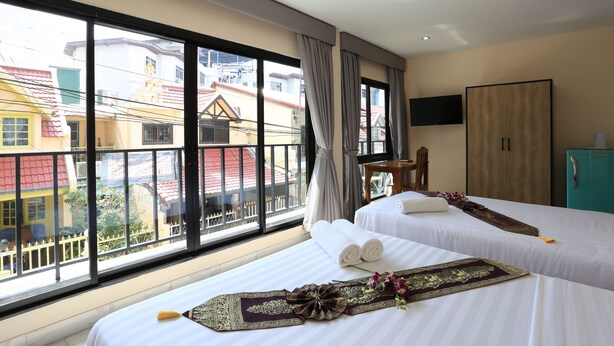 A hotel room with two beds, a large window with a view of colorful buildings, a wooden wardrobe, and a flat-screen TV on the wall. - The Siam Guest House (Pattaya)