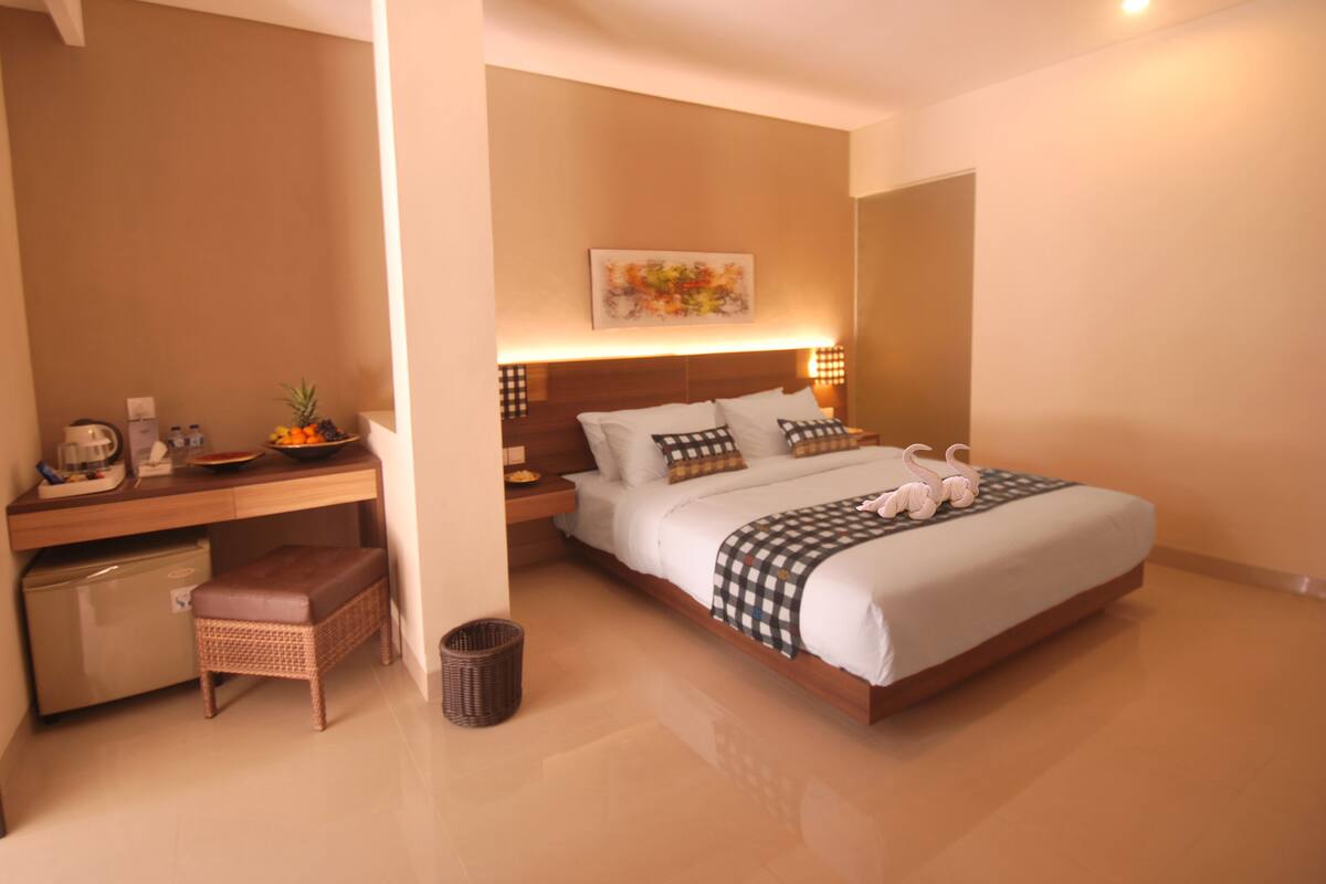 A modern hotel room with a large bed, a desk with a computer, a small kitchenette, and a trash bin.