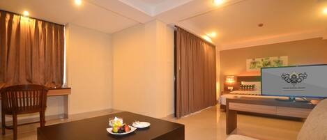 Una habitación de hotel con cama, televisión, mesa de centro con fruta, una silla y cortinas.