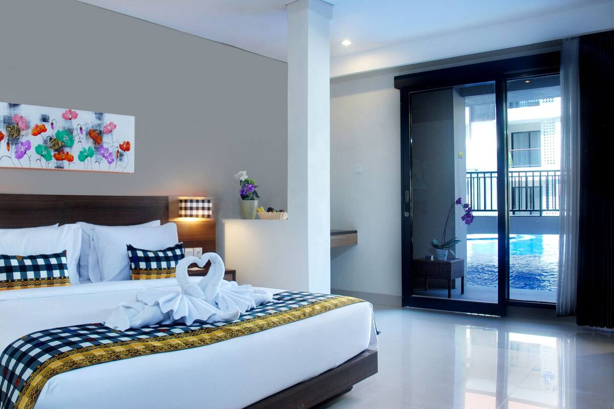 A modern hotel room with a large bed, a painting on the wall, a nightstand with a phone, and a view of a pool through the glass door.