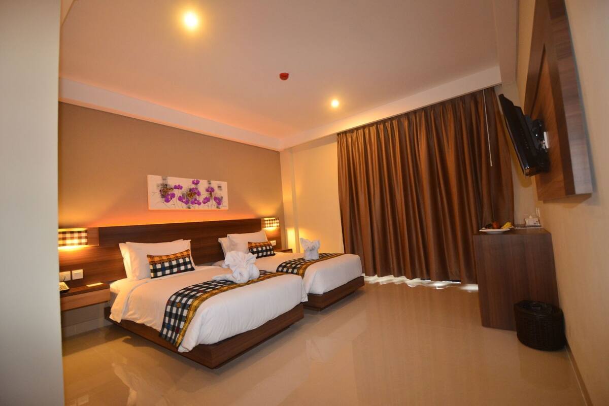 A hotel room with two beds, a flat-screen TV, a painting, and a large window with curtains.