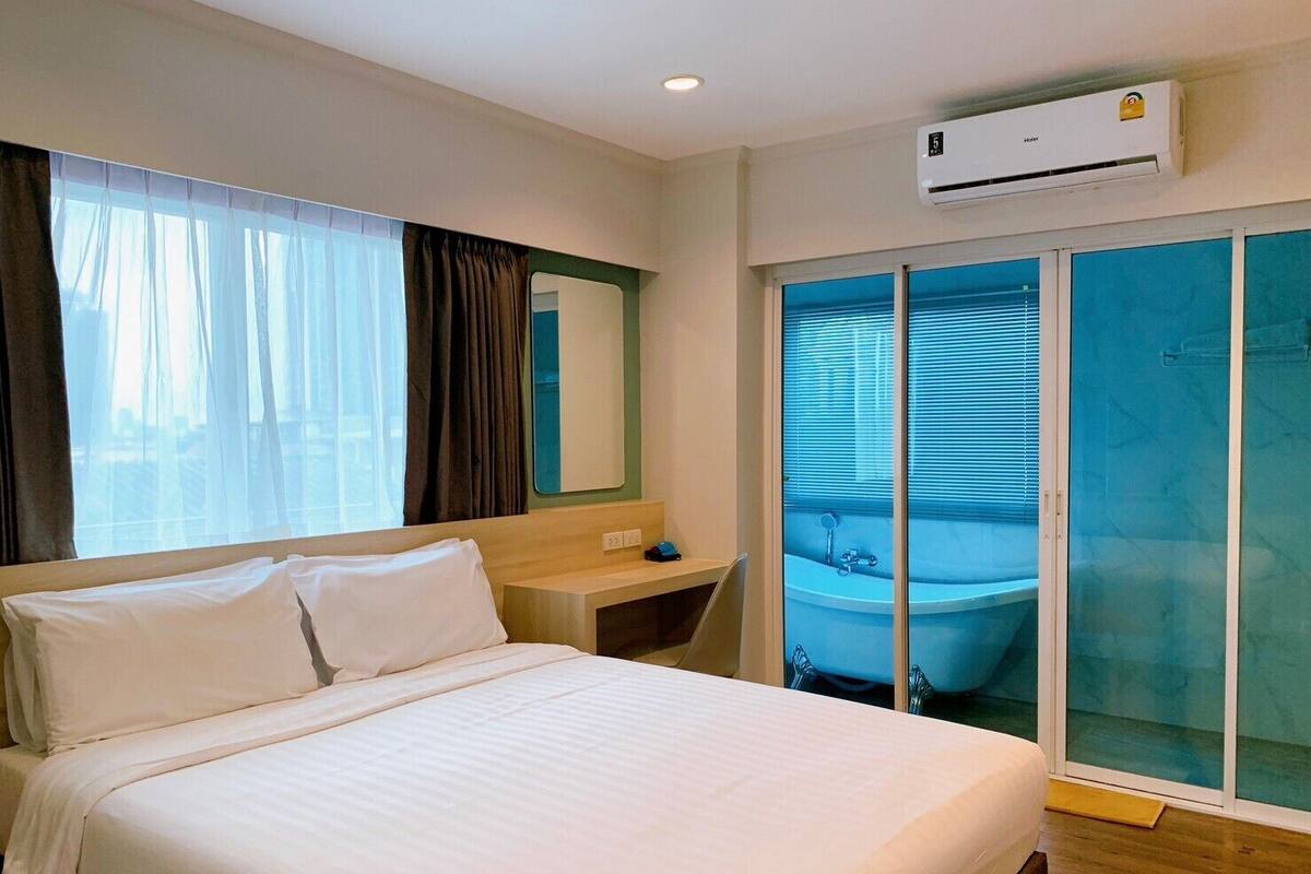 Hotelkamer met een bed, een klein nachtkastje, een spiegel, een airconditioningsysteem en een schuifglazen deur naar een badkamer met een bad.