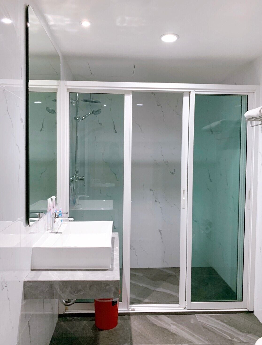 A modern bathroom with a white sink, glass shower enclosure, and a red bucket.