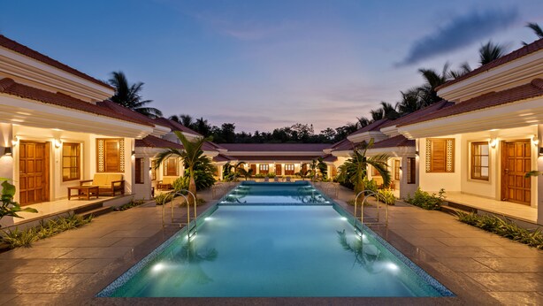 A swimming pool with a tiled deck, surrounded by two-story buildings with wooden doors and windows, and palm trees. - Leoney Resort (Vagator)