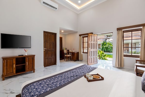 A hotel room with a large bed, a flat-screen TV, a wooden cabinet, and a view of the garden through the open door. - Leoney Resort (Vagator)