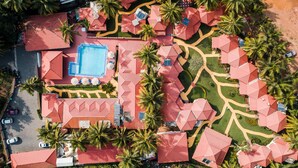 An aerial view of a resort with a central swimming pool, multiple red-roofed buildings, and palm trees. - Leoney Resort (Vagator)