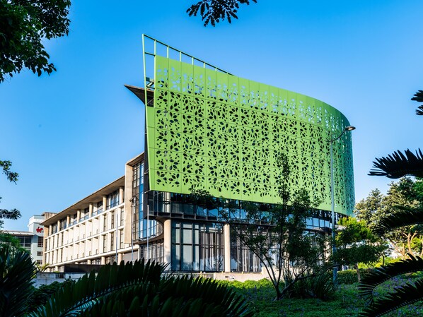 Un edificio moderno con una fachada verde perforada y un entorno de abundante vegetación y árboles.