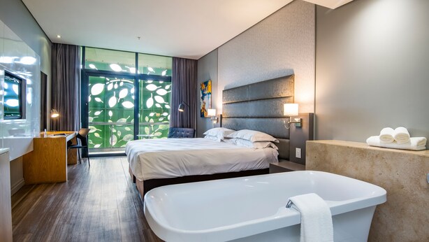 A modern hotel room with a large bed, a sitting area, a bathtub, and a view through the window. - aha Gateway Hotel (Umhlanga)