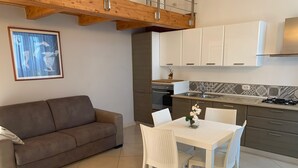 Un espace de vie compact comprenant un canapé marron, une table à manger blanche avec des chaises, et un coin cuisine avec des meubles blancs et un dosseret carrelé.
