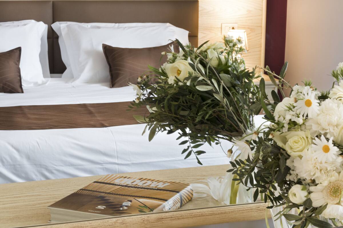 Een hotelkamer met een net opgemaakt bed, een nachtkastje met een boek en een bloevas, en een wandlamp.