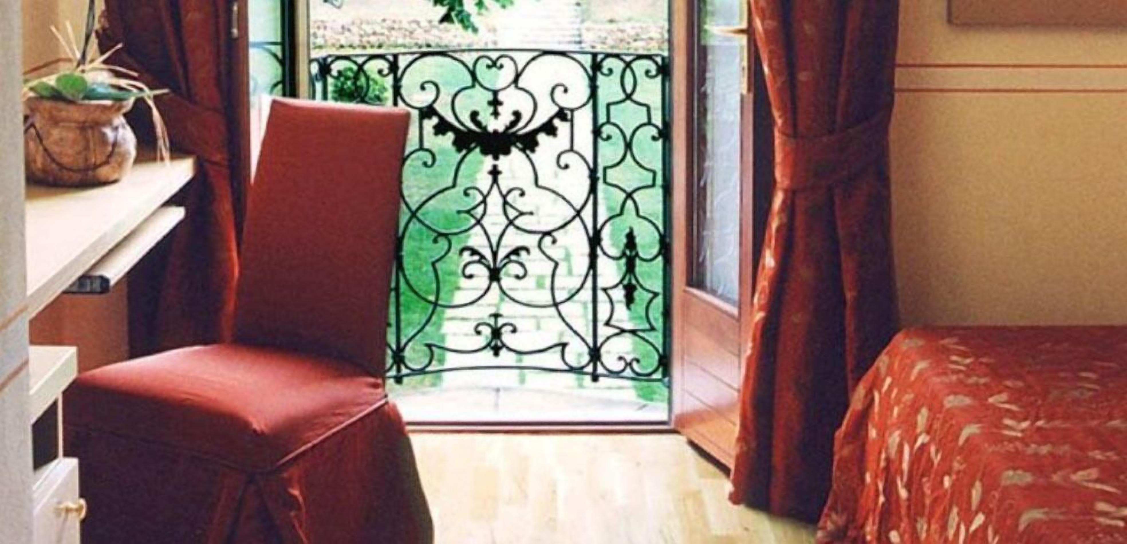 A room with a red chair, a bed with a patterned bedspread, a window with a decorative iron gate, and a potted plant on a shelf.