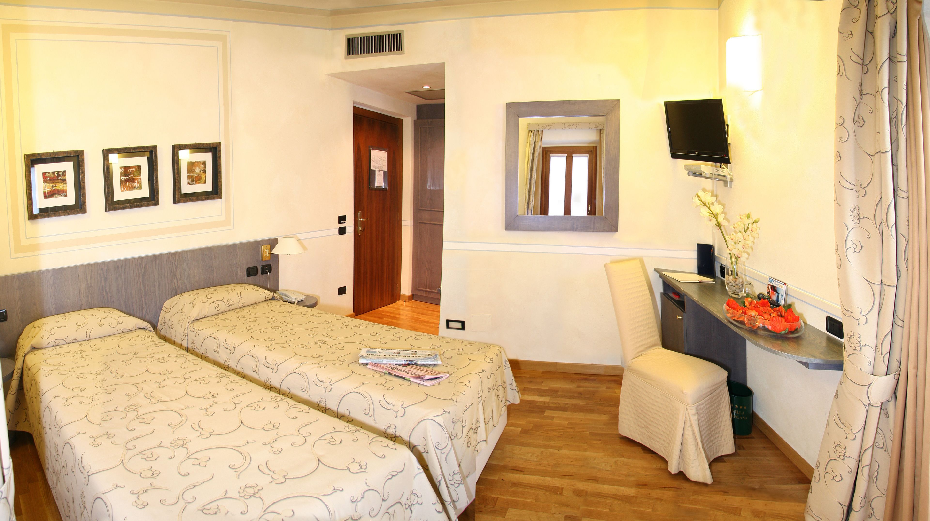 Ein Hotelzimmer mit zwei Betten, einem Schreibtisch mit Stuhl, einem Fernseher und gerahmten Bildern an der Wand.