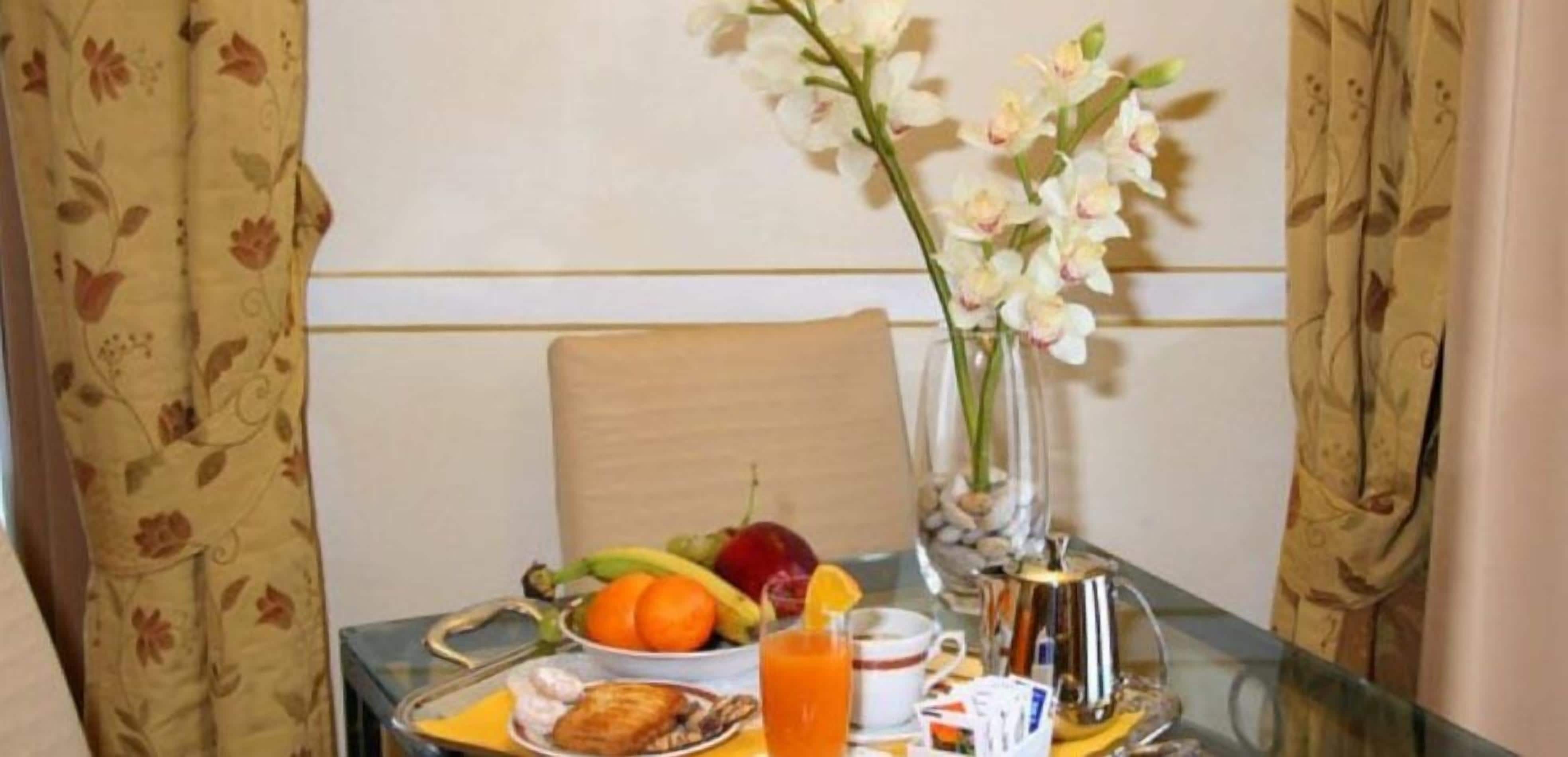 Disposição de café da manhã com frutas, um copo de suco de laranja e um vaso de flores sobre uma mesa.