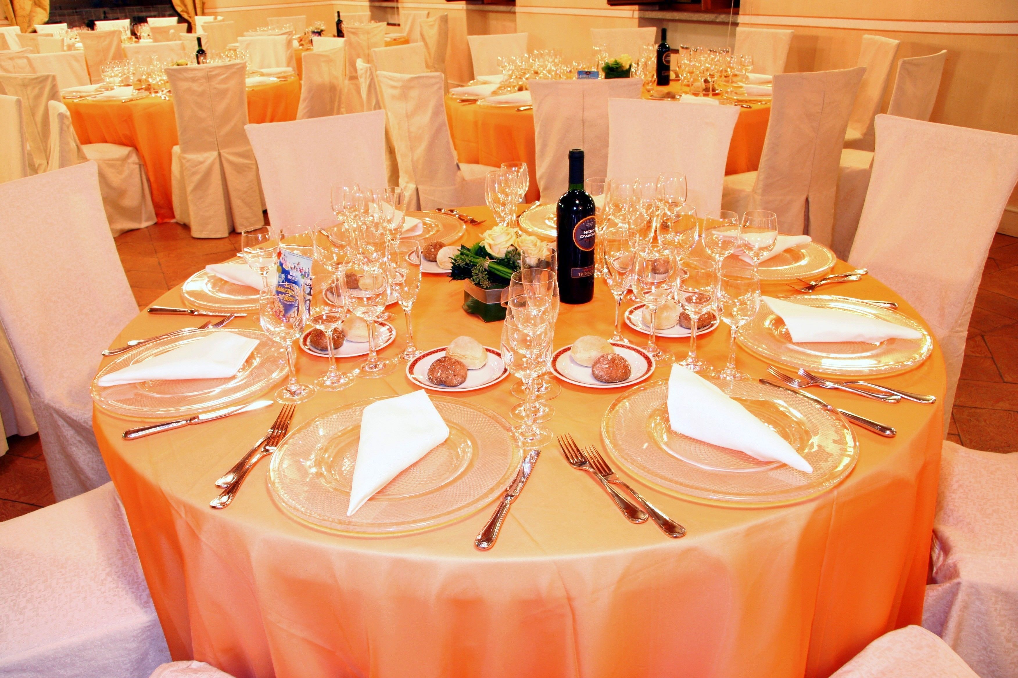 A banquet hall with round tables set for a formal event, complete with place settings, wine glasses, and a bottle of wine.