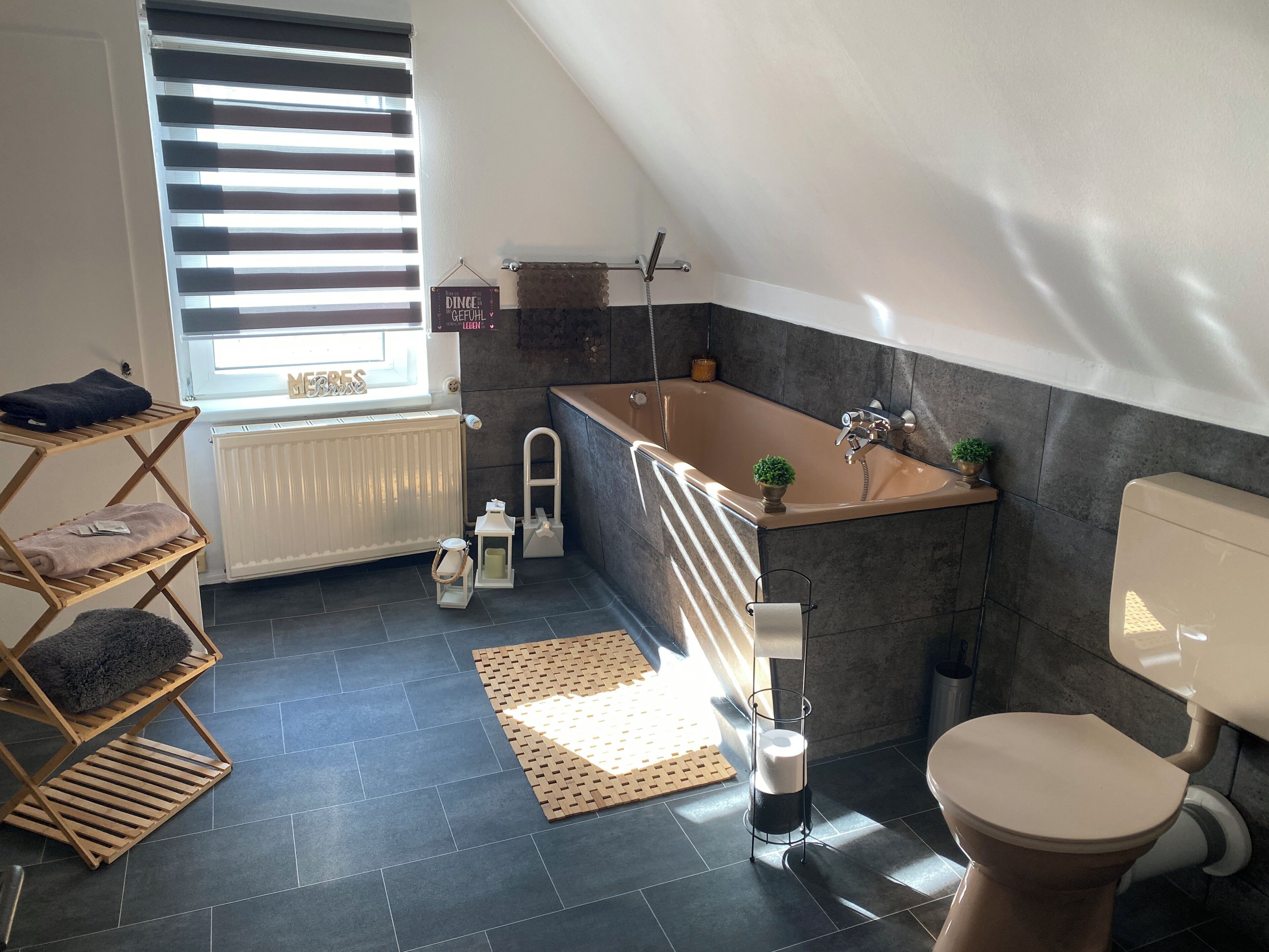 Ein modernes Badezimmer mit einer freistehenden Badewanne, einer Toilette und einem Fenster mit Jalousien.