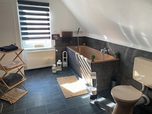 A modern bathroom with a freestanding bathtub, a toilet, and a window with blinds. - Altstadt Hotel Gosequell (Goslar)