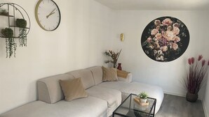 Una sala de estar moderna con un sofá color beige, una mesa de centro de vidrio, un reloj de pared redondo, un cuadro floral y una planta en maceta.