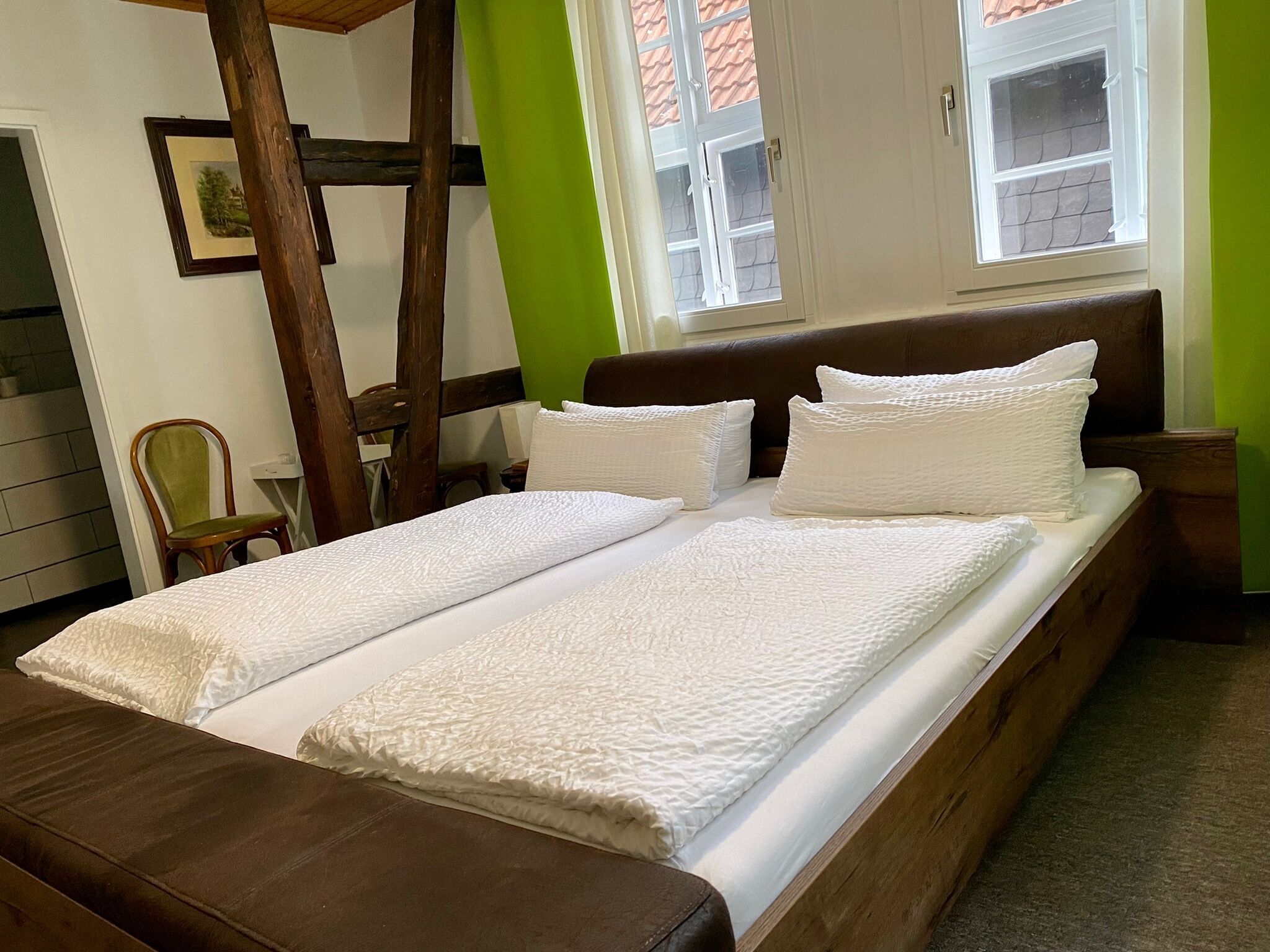 Cama doble con ropa blanca, cabecera de madera y ventana con vista a un edificio.