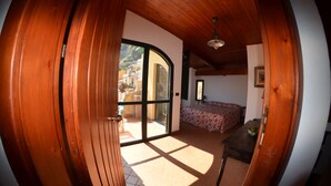 A room with a wooden ceiling, a bed with a floral bedspread, a table with a bowl, and a view of a courtyard through a glass door. - Le Piccole Grotte (Scilla)