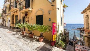 A narrow street with buildings on both sides, potted plants, and a clear view of the sea. - Le Piccole Grotte (Scilla)