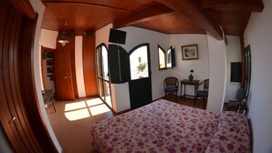 A bedroom with a wooden ceiling, a bed with a floral bedspread, a desk with a chair, and a window with a view of the outside. - Le Piccole Grotte (Scilla)