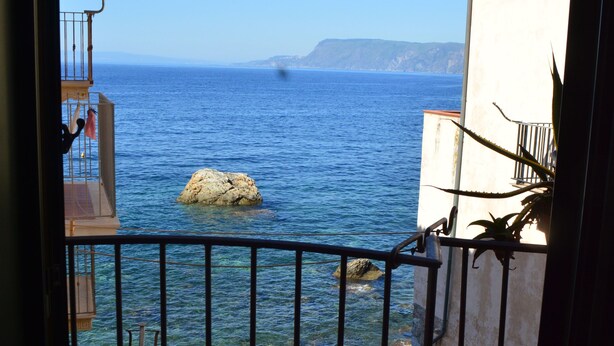 A balcony with two cushioned chairs, a railing, and a view of the sea and distant mountains. - Le Piccole Grotte (Scilla)