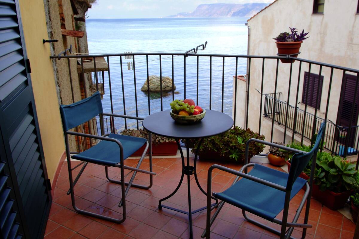 Een balkon met twee blauwe stoelen, een klein rond tafeltje met een fruitschaal en potplanten.