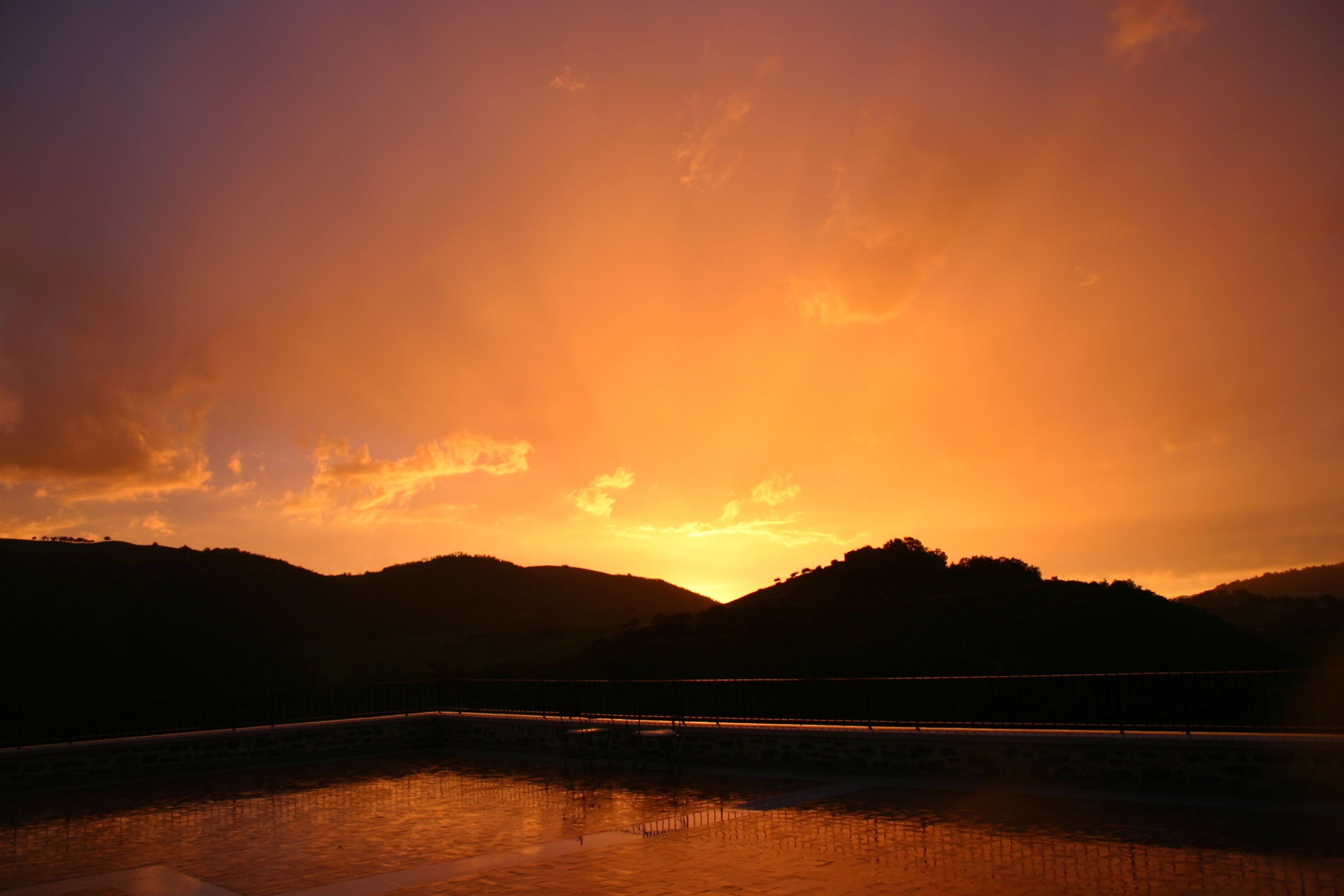 A sunset over a hilly landscape with a bridge and a body of water reflecting the sky.